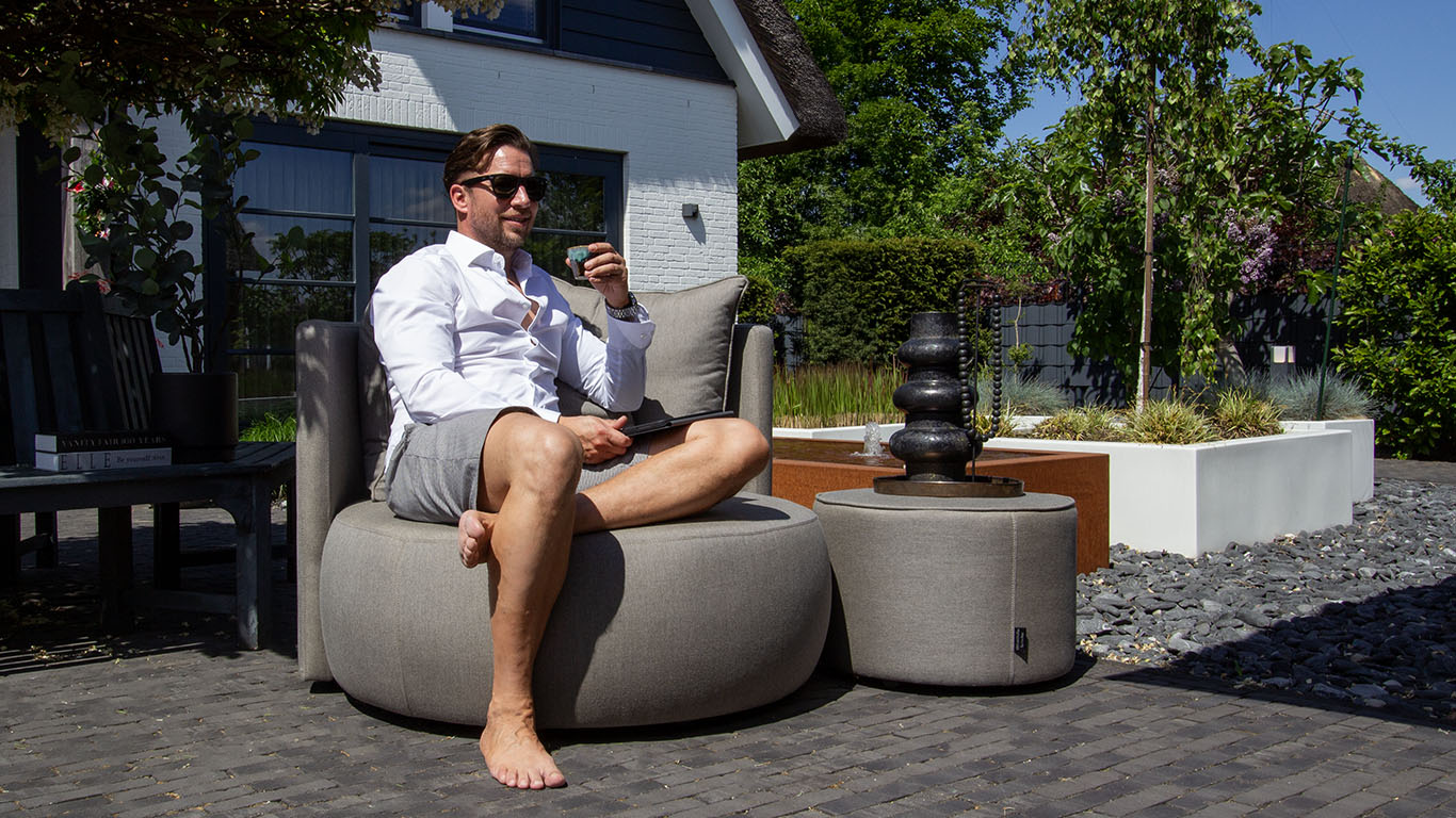 UrbanSofa Outdoor Fauteuil Ibiza in Natte Grey Chine met Swing Functie en Bellagio Outdoor Poef low in Natte Grey Chine kop koffie drinken in de Tuin 1